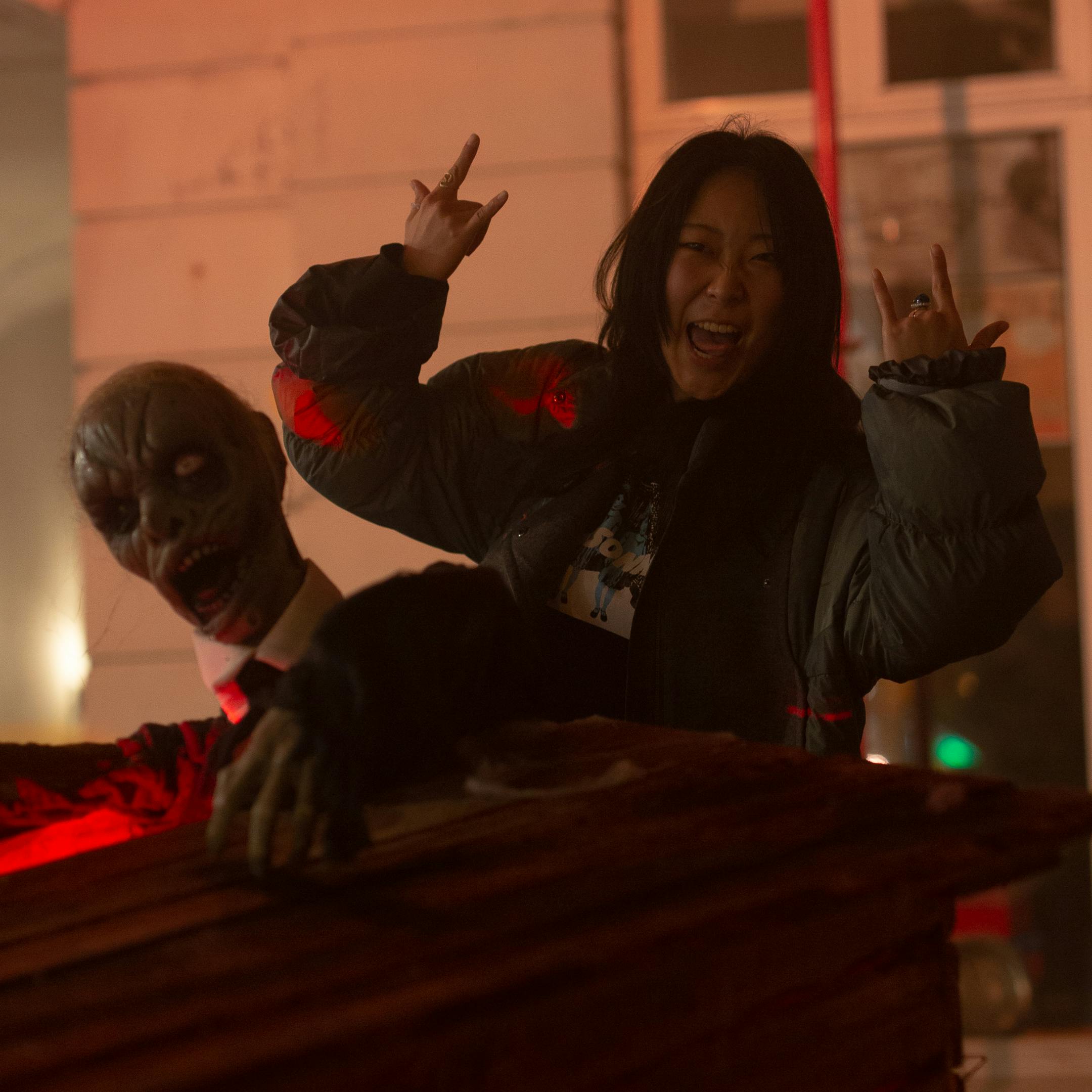 Asian woman celebrates Halloween with spooky zombie figure indoors, exuding happiness and excitement.