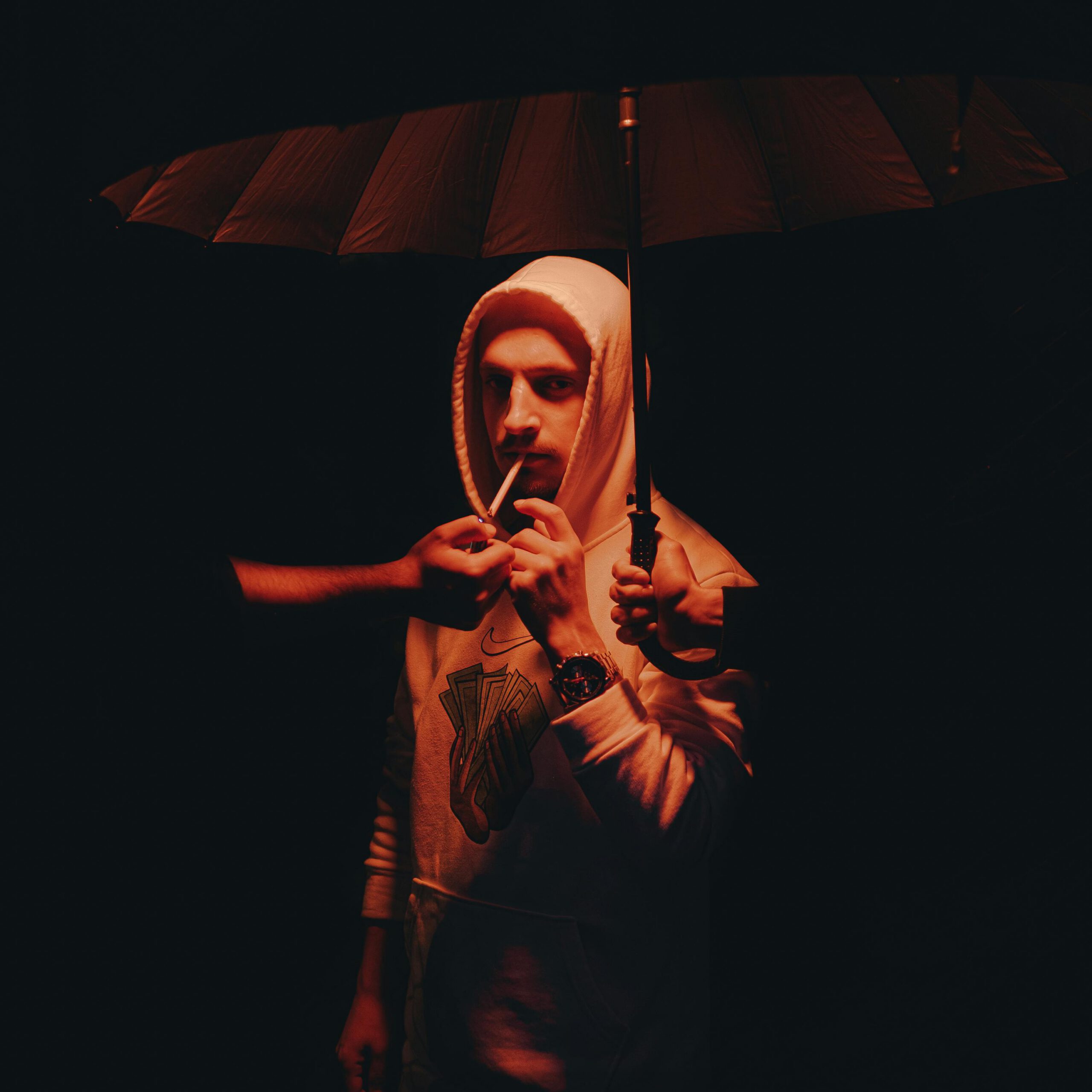 A man in a hoodie smokes under an umbrella, lit by dramatic red light.