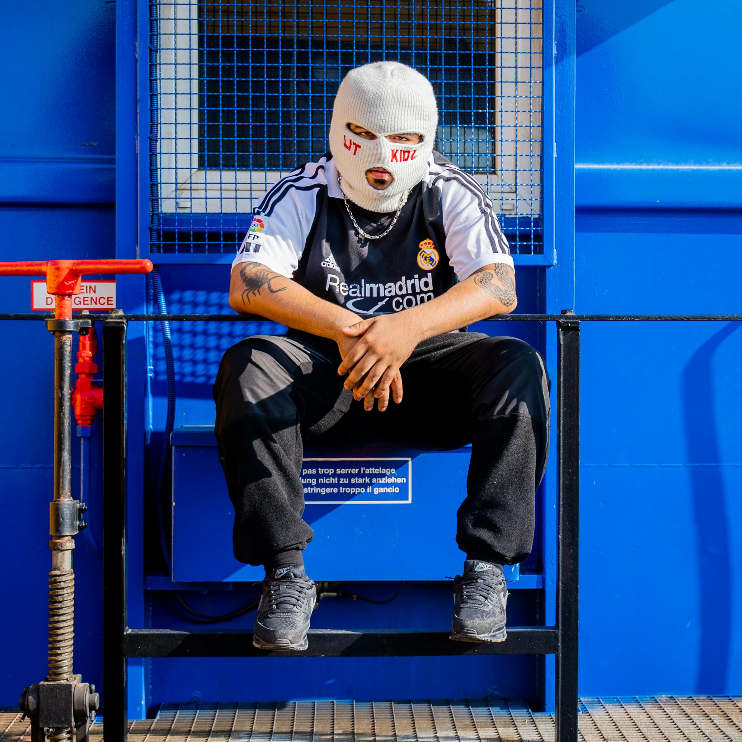 Person in a Real Madrid jersey wearing a ski mask, posing in an urban setting.